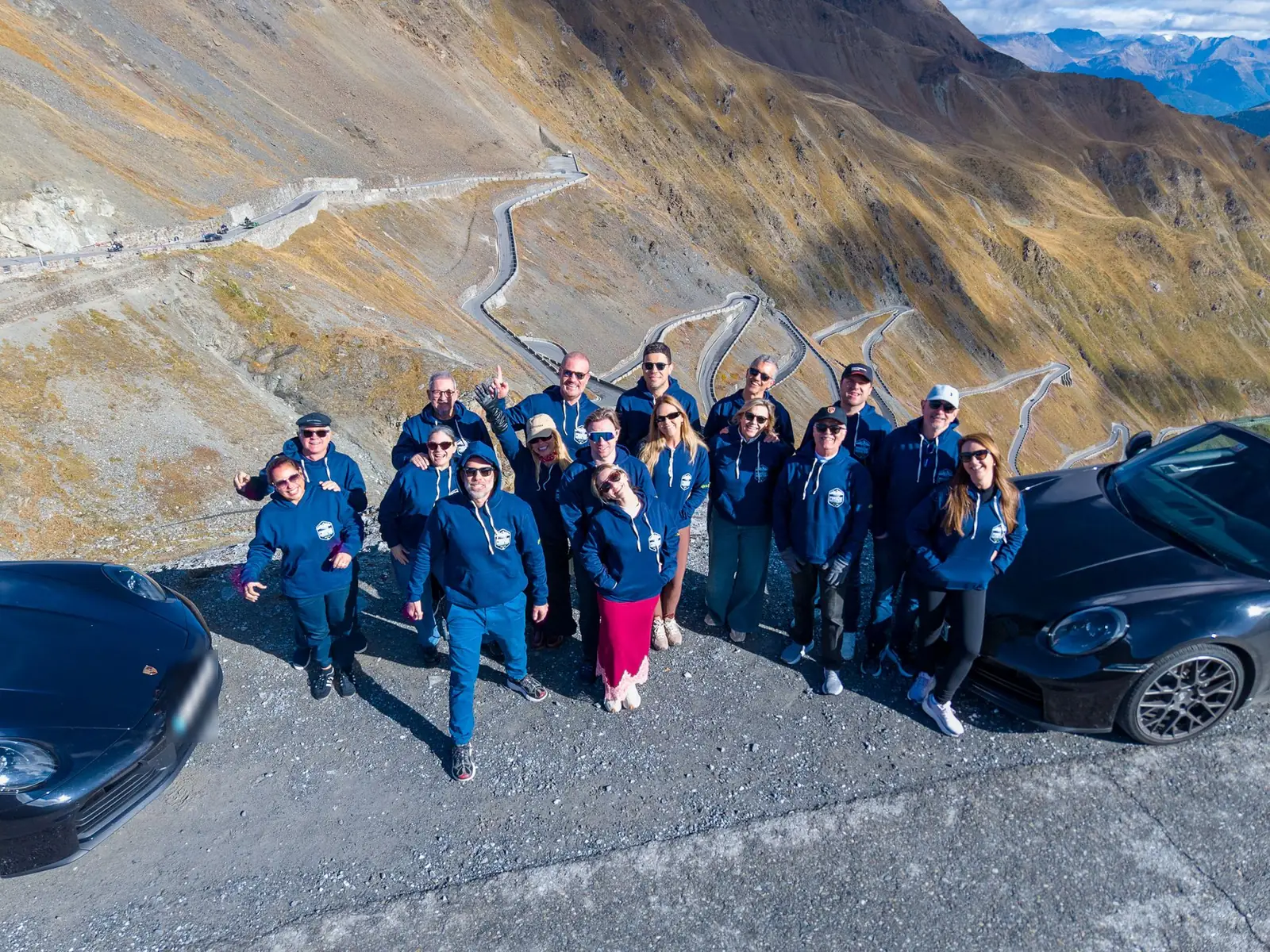 Grupo do Porsche Driving Tour Legends of the Alps da ROXMOTORS no alto do Passo Stelvio, com as curvas em zigue-zague ao fundo, na Itália
