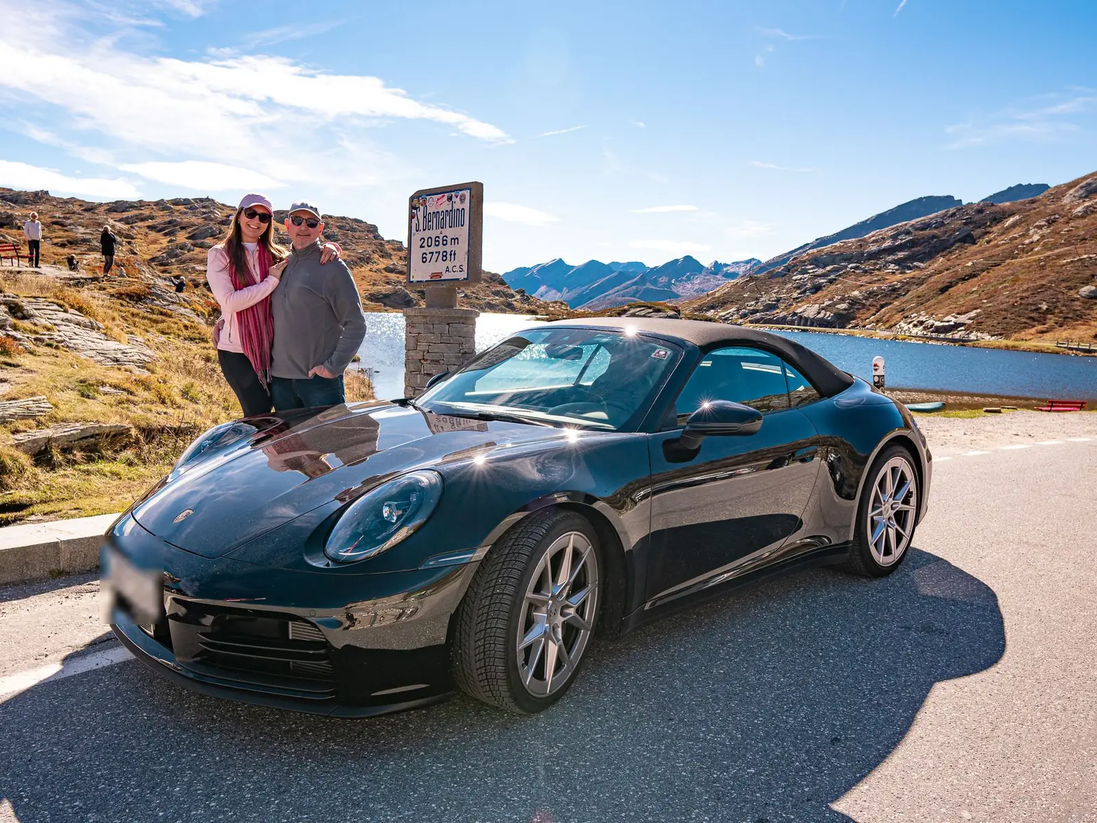 Casal de participantes com Porsche 911 Cabriolet preto junto à placa do Passo San Bernardino, a 2.066 metros, na Suíça