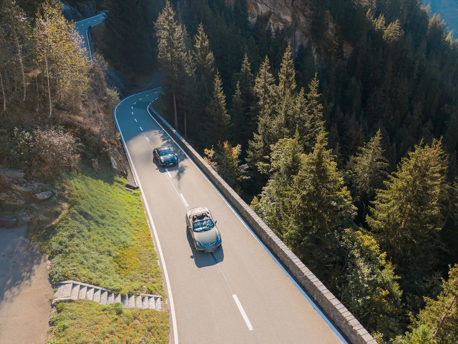 Dois Porsche conversíveis em estrada de montanha entre pinheiros nos Alpes, vistos de drone